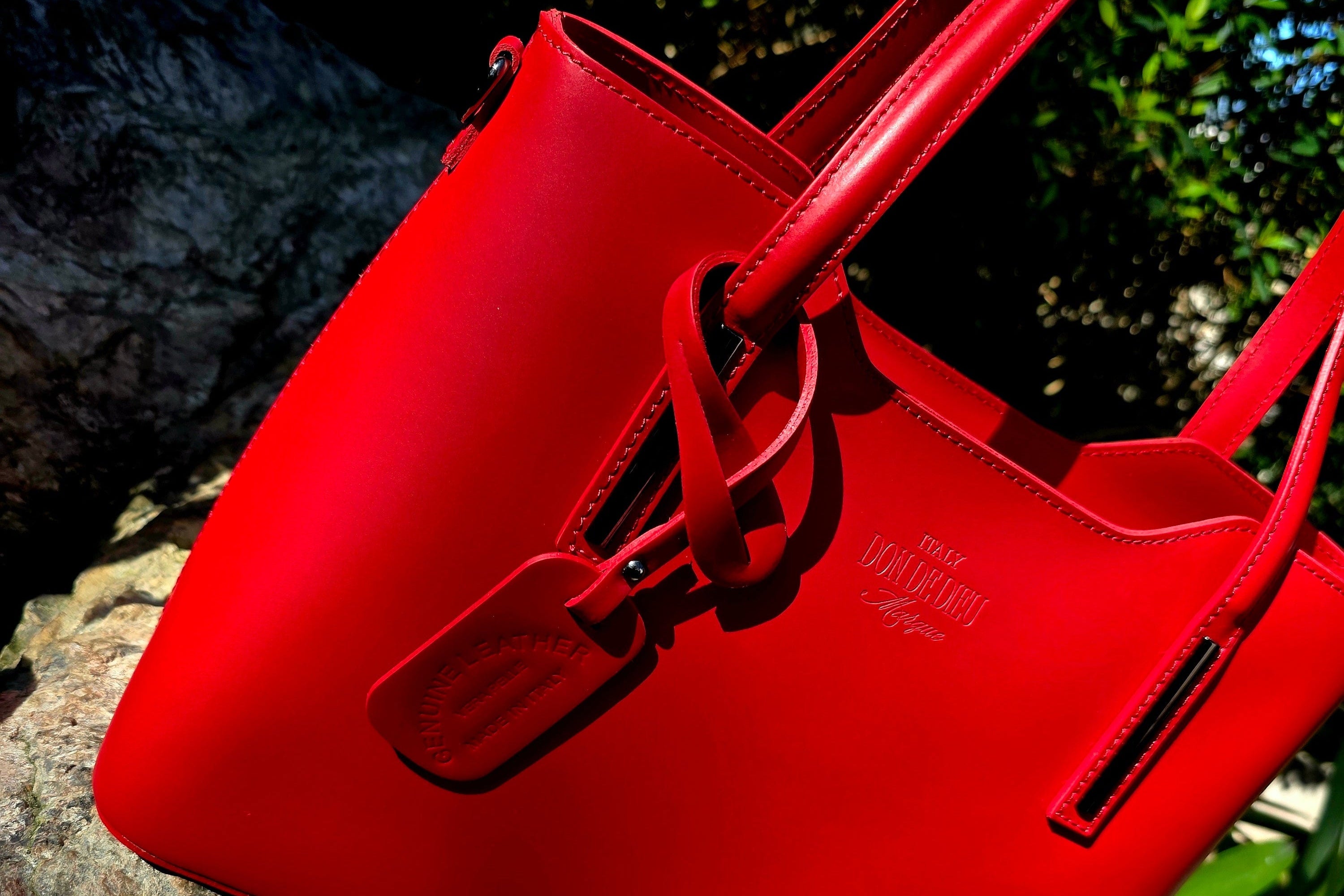 Side view of Red Italian handbag with a brand logo on a stone surface with greenery in the background. - Don de Dieu Marque