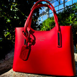 Full view of Red Italian handbag with a visible brand logo on a stone surface with greenery in the background. - Don de Dieu Marque