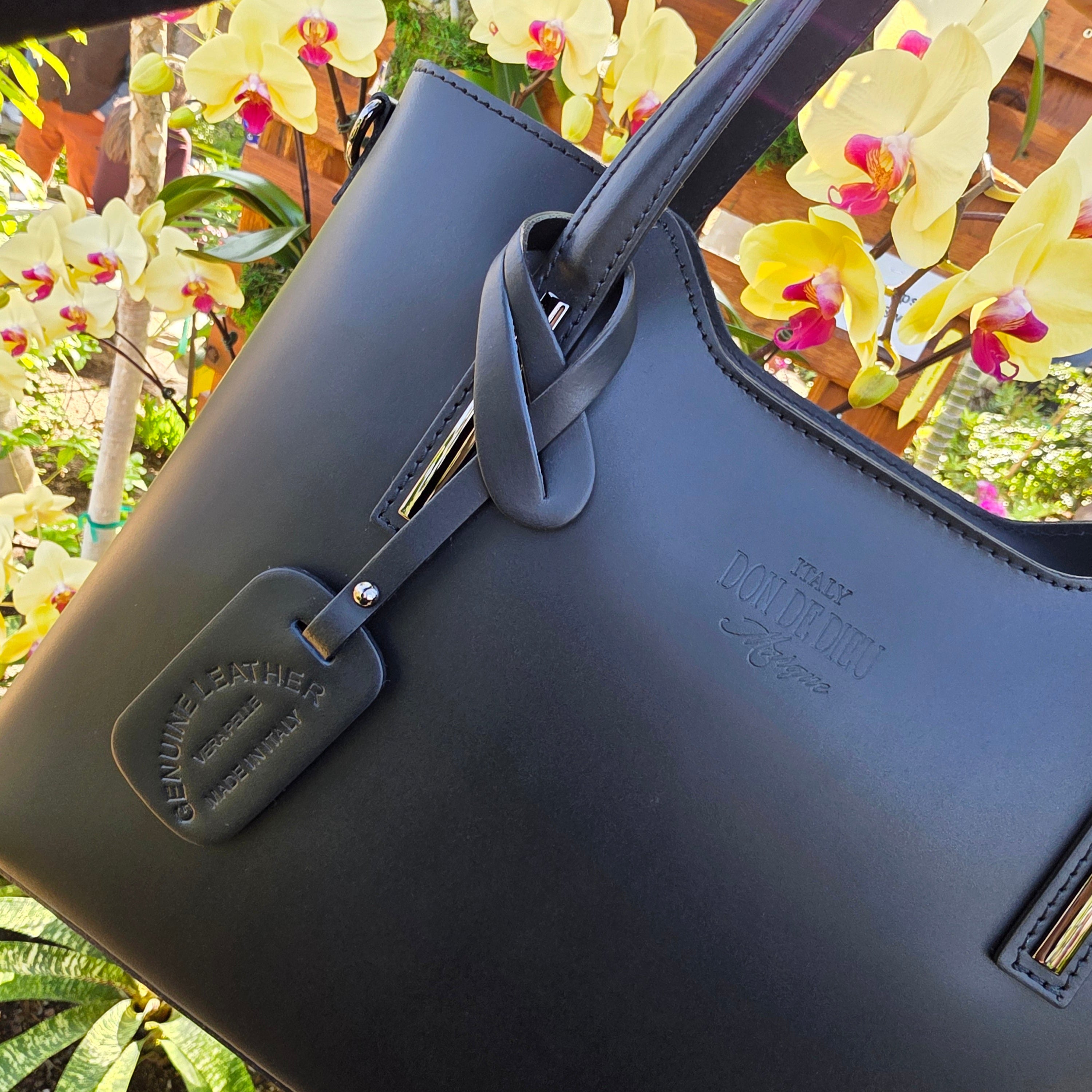 Black Italian handbag with visible brand logo being held against a real yellow orchid blurred floral background. - Don de Dieu Marque
