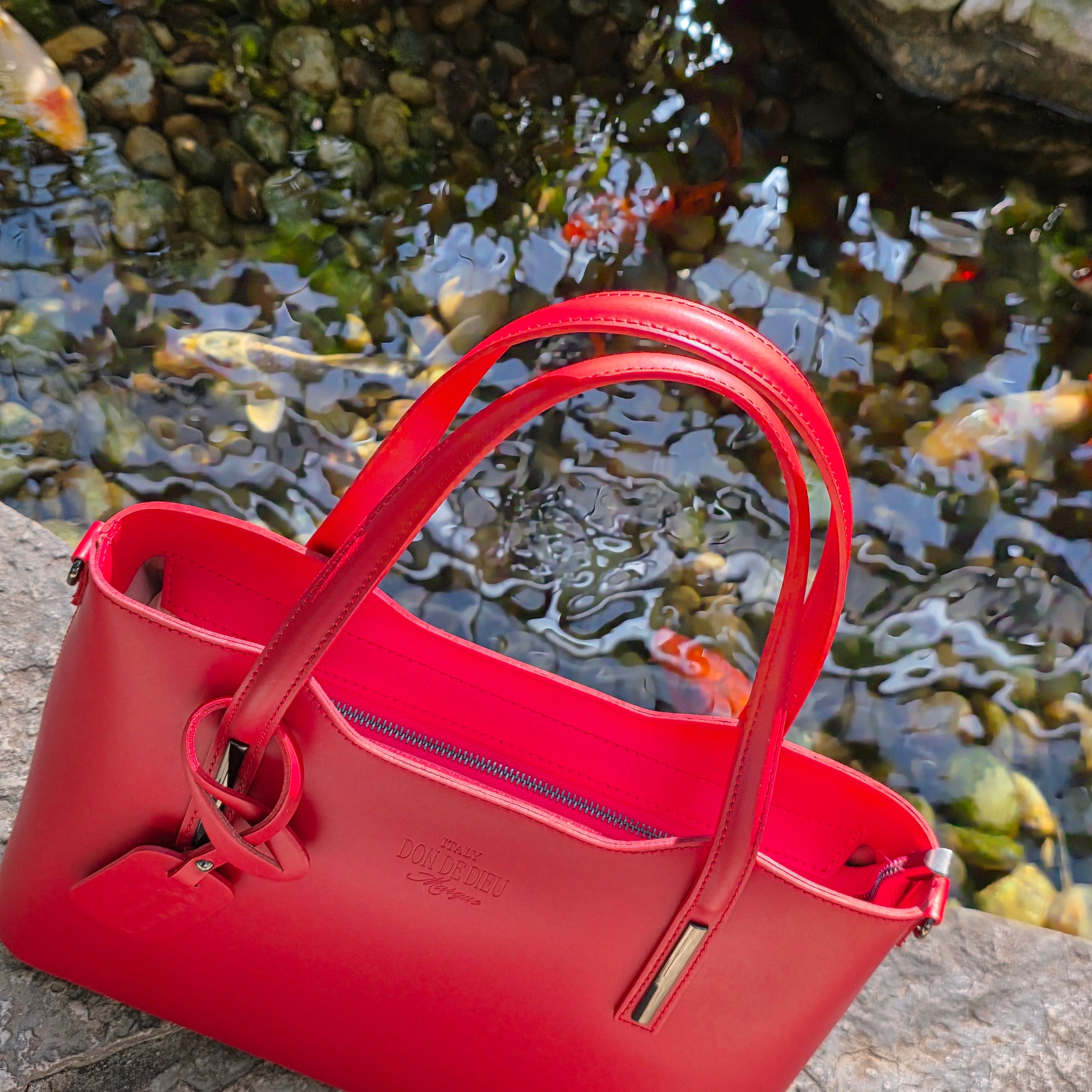 Red Italian leather handbag leaning against a stone wall with water and Koi fish in the background. - Don de Dieu Marque