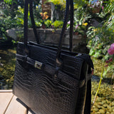Side angle of Black Croc Italian leather handbag with a silver clasp on a wooden surface with a garden background. - Don de Dieu Marque 