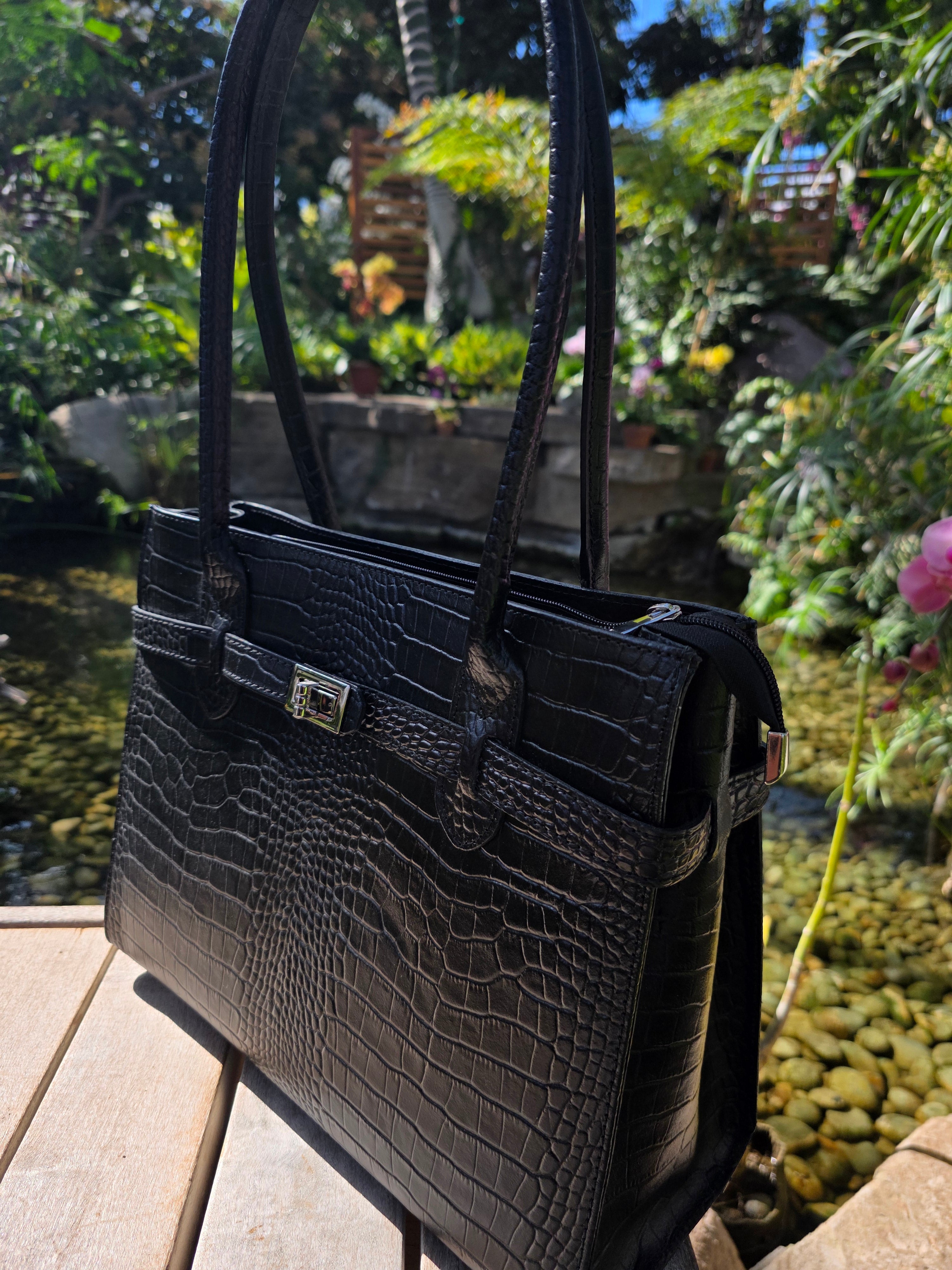 Side angle of Black Croc Italian leather handbag with a silver clasp on a wooden surface with a garden background. - Don de Dieu Marque 