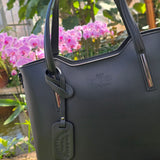 Authentic Black Italian handmade bag with visible brand logo against a blurred natural flower background. - Don de Dieu Marque