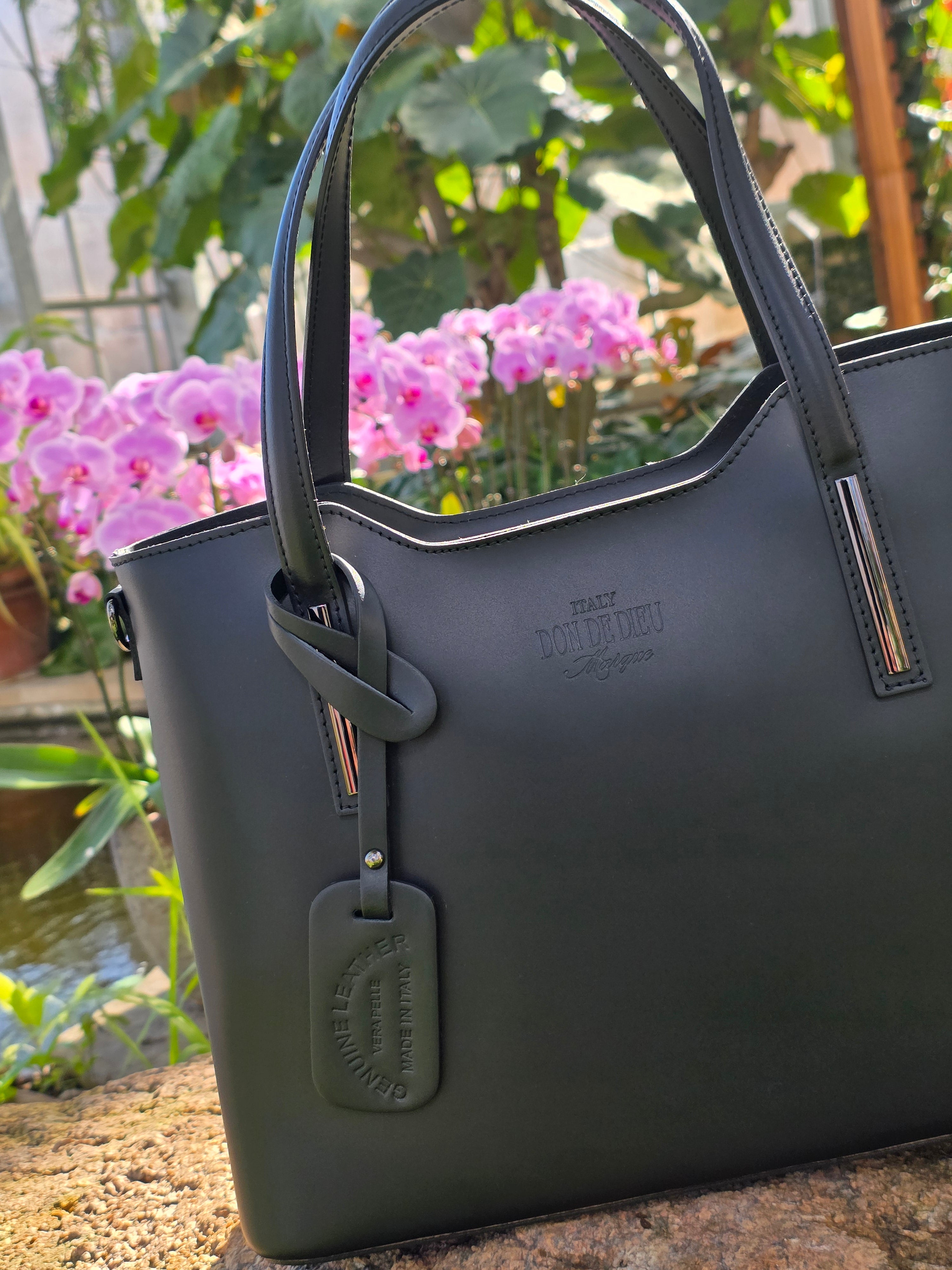 Authentic Black Italian handmade bag with visible brand logo against a blurred natural flower background. - Don de Dieu Marque