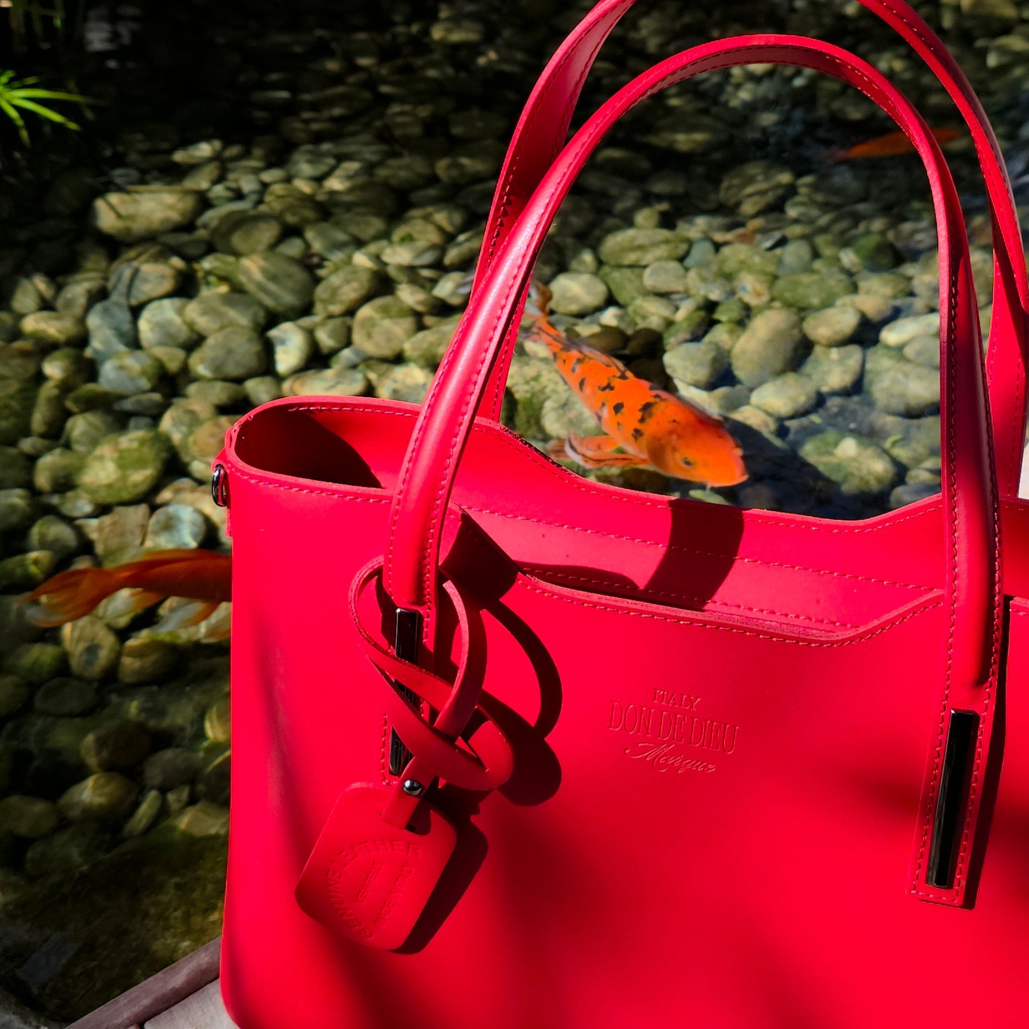 Up close view of Red Italian handbag with a brand logo on a wooden deck with a pond in the background. - Don de Dieu Marque