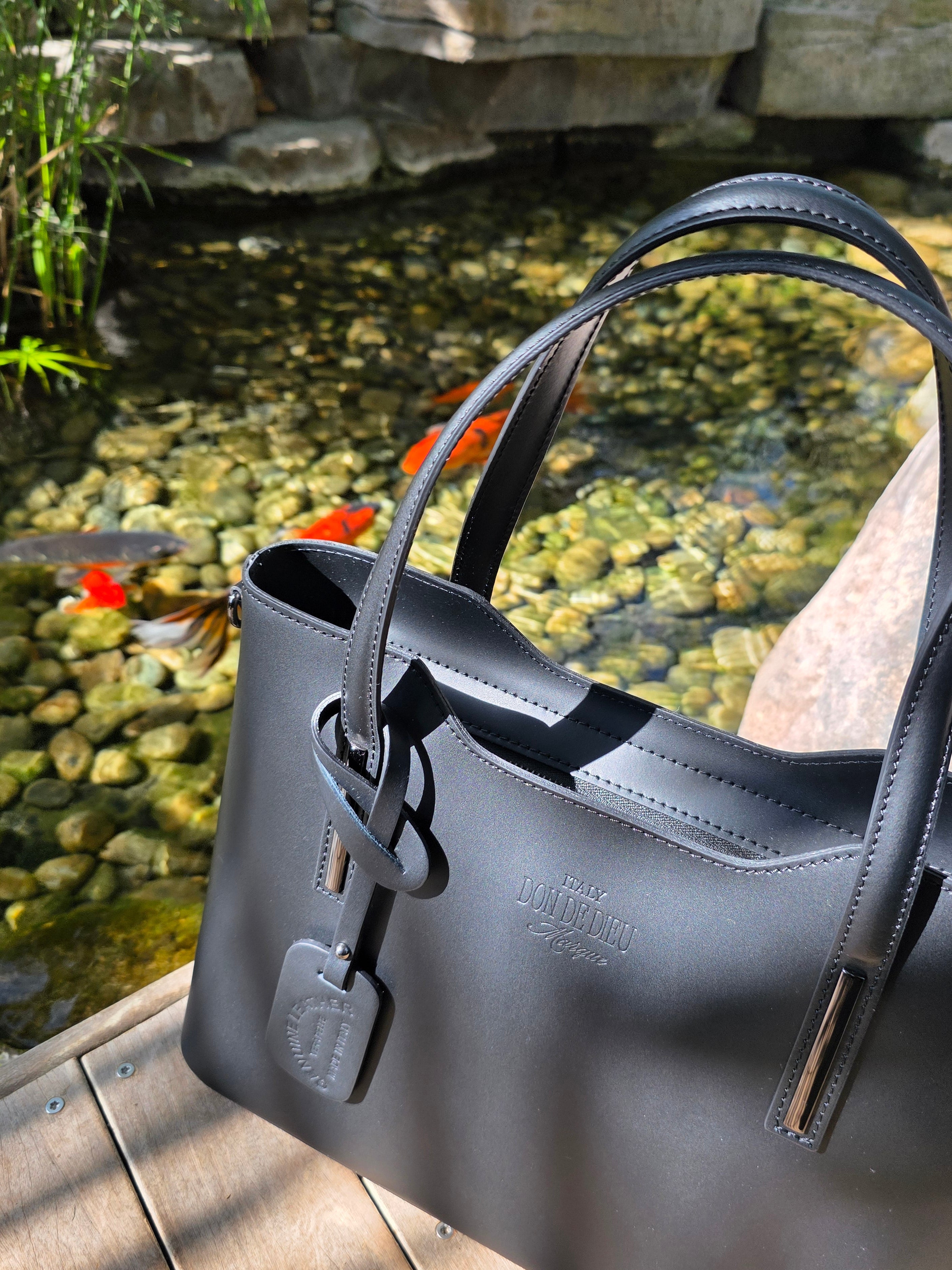 Black Italian leather bag with black strap against a natural Koi fishpond with stones background. - Don de Dieu Marque