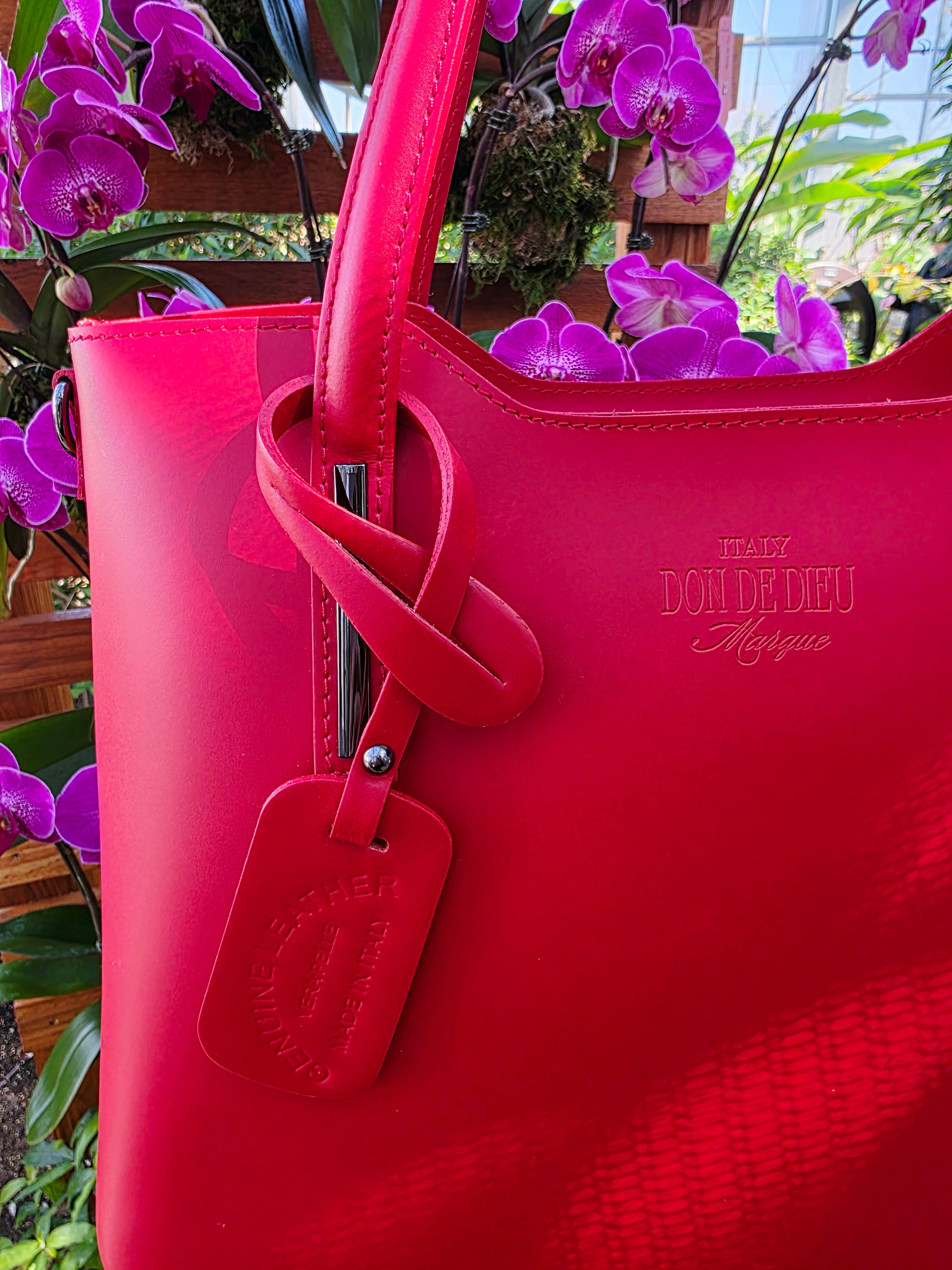 Red Italian leather handbag with visible brand logo that reads, "Italy Don de Dieu Marque" against a background of purple flowers