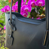 Side view of Black Italian handbag with a brand logo in front of purple flowers. -  Don de Dieu Marque