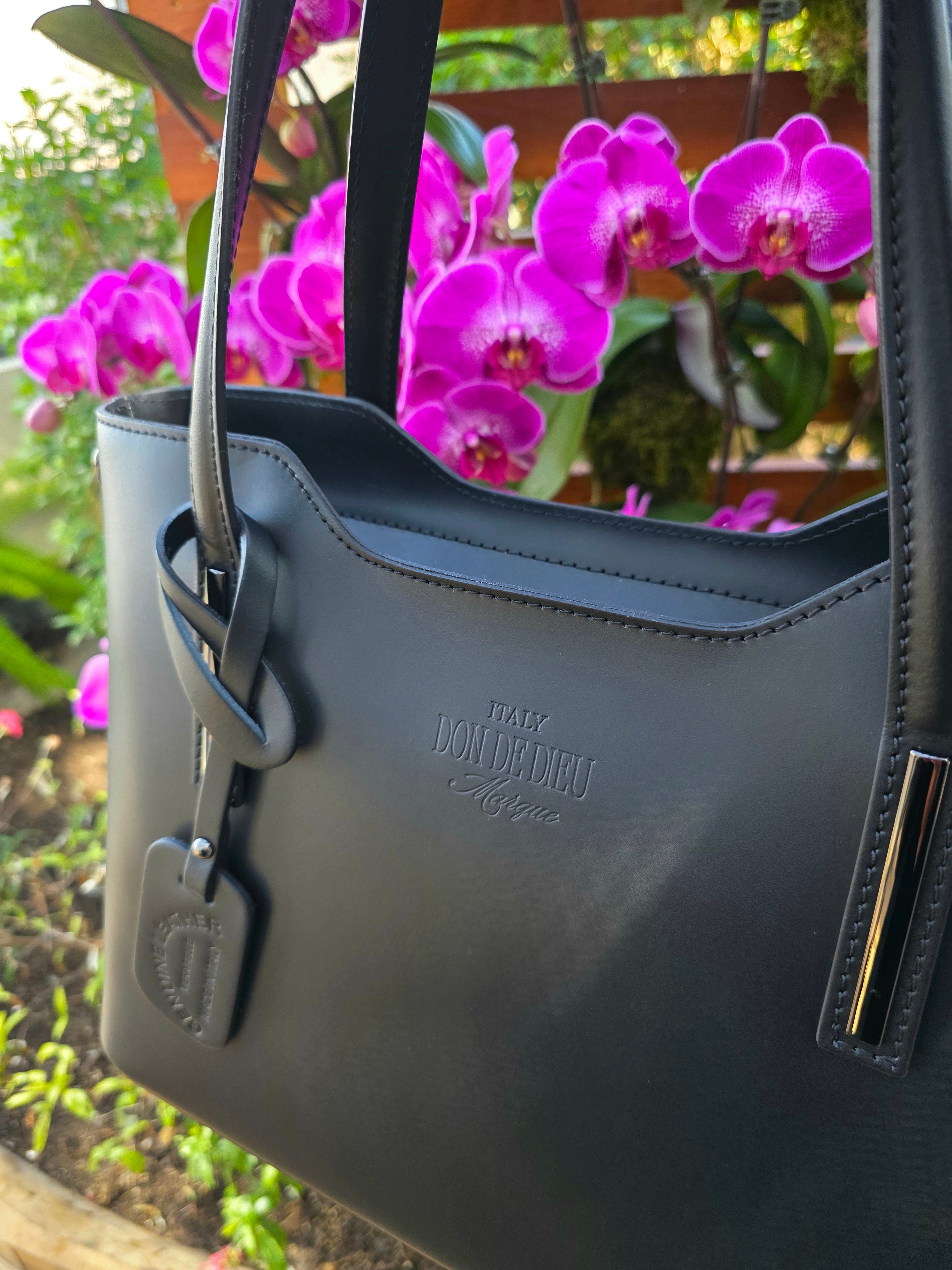 Side view of Black Italian handbag with a brand logo in front of purple flowers. -  Don de Dieu Marque