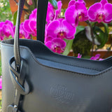 Cropped up close Black Italian handbag with detailed stitching of brand logo in front of purple orchid flowers. - Don de Dieu Marque