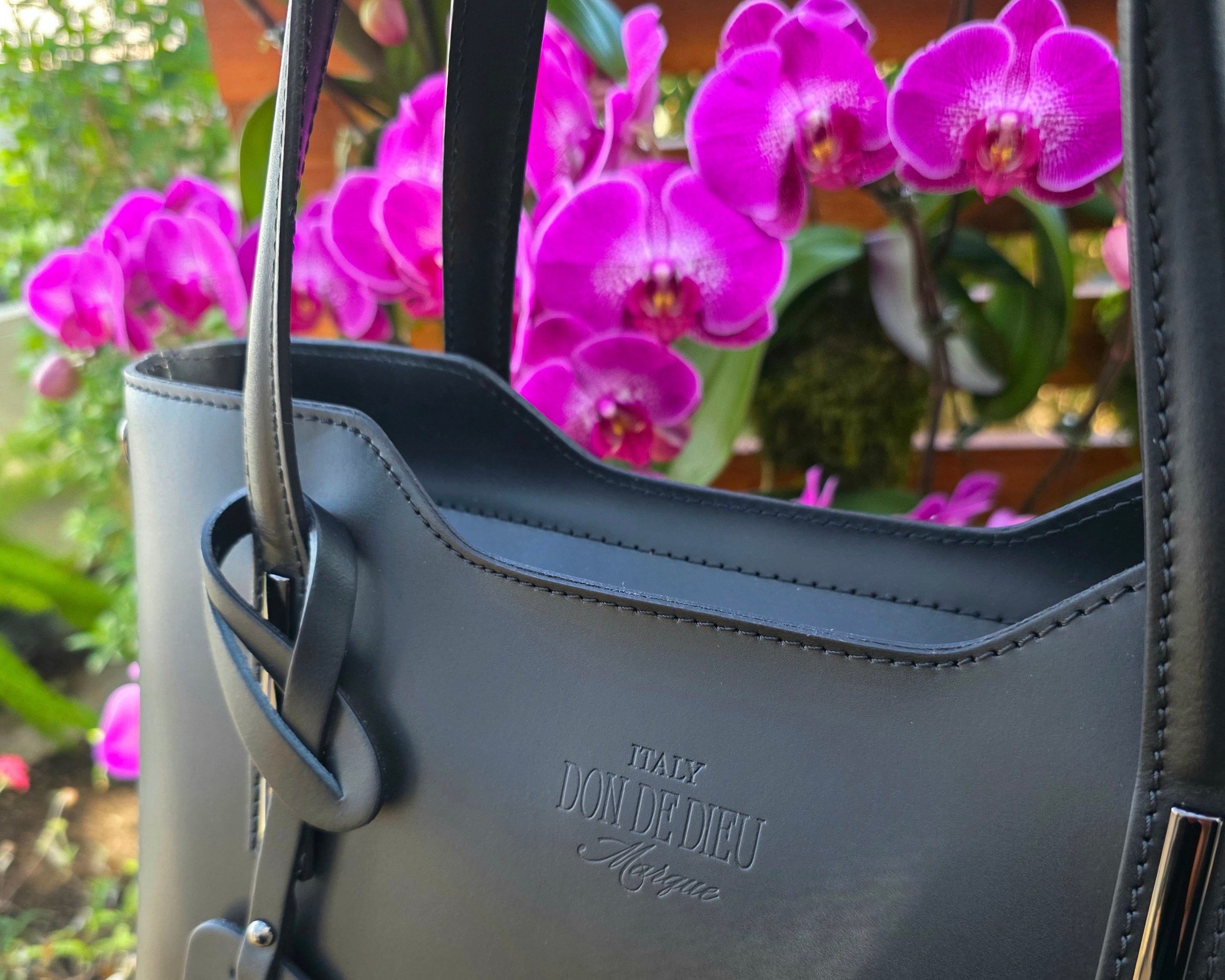 Cropped up close Black Italian handbag with detailed stitching of brand logo in front of purple orchid flowers. - Don de Dieu Marque