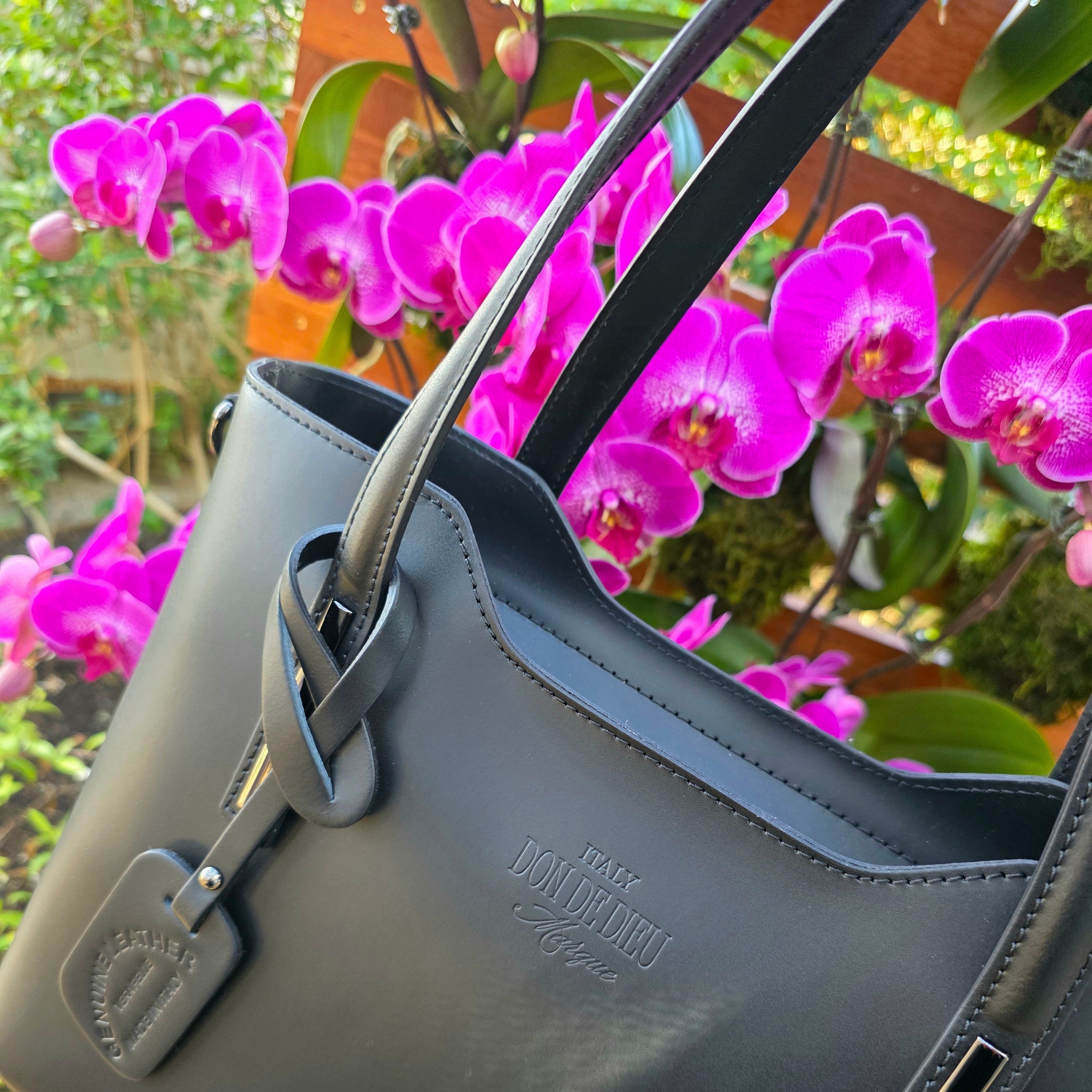 Beautiful front side of Black Italian handbag with visible brand logo in front of pink orchids. - Don de Dieu Marque