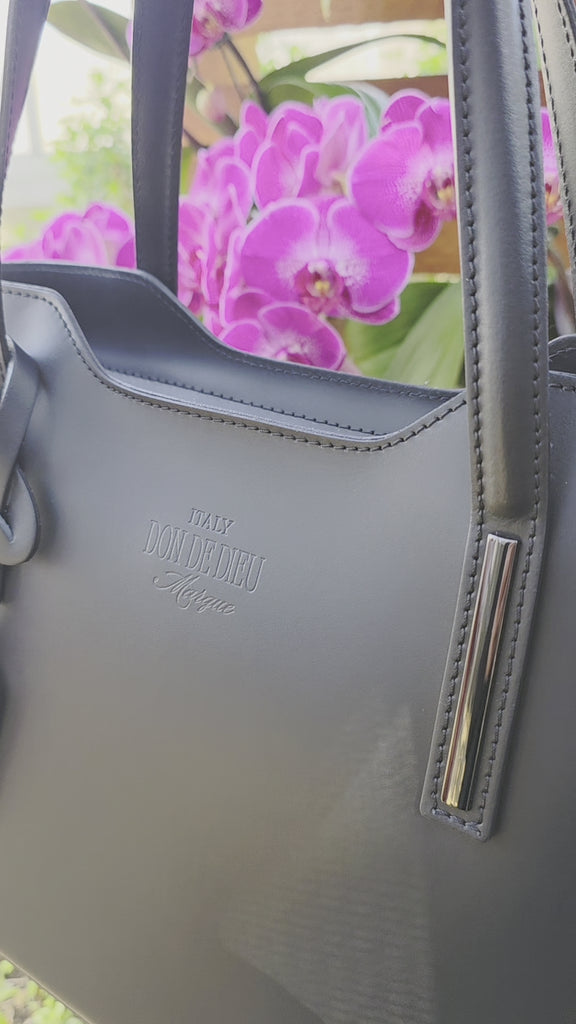 Black Italian leather handbag with brand logo up close video to capture details with purple orchid flowers and greenery background. - Don de Dieu Marque